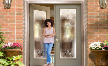 woman stands in partially open doorway: two gray ProVia doors with large decorative windows on a brick house with flower pots