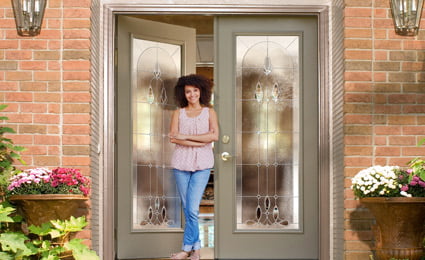 ProViaDoors_Legacy_425x260 woman stands in partially open doorway: two gray ProVia doors with large decorative windows on a brick house with flower pots