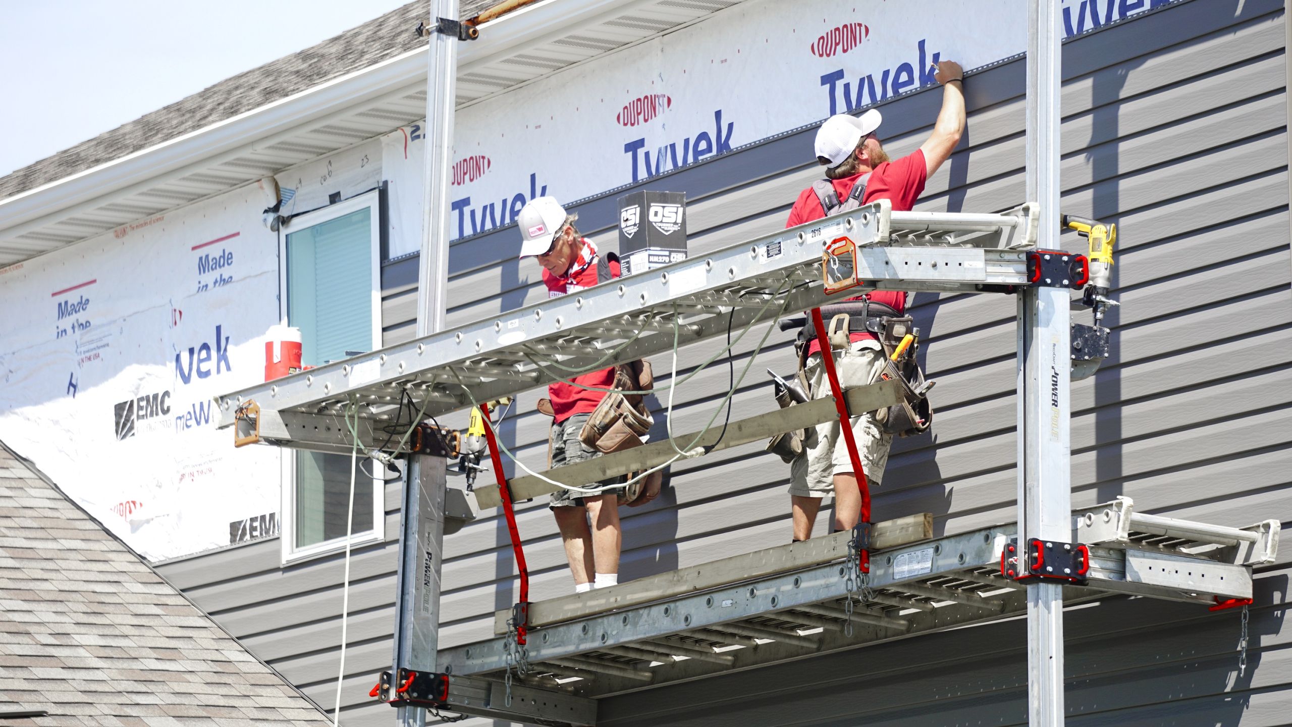Two installers on a raised platform attaching gray seamless steel siding panels to a house exterior.