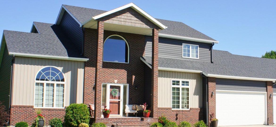 Front view of a two-story house with dark gray asphalt shingle roof, brick and beige siding, multiple large windows, and a white double garage door under a clear blue sky.