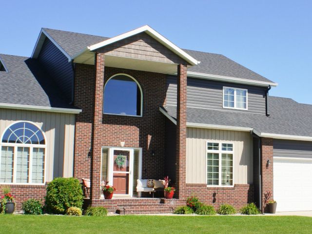 Front view of a two-story house with dark gray asphalt shingle roof, brick and beige siding, multiple large windows, and a white double garage door under a clear blue sky.