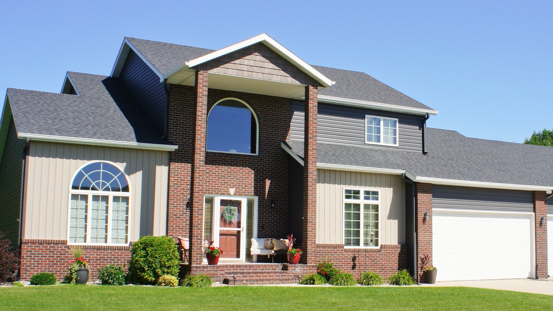 Front view of a two-story house with dark gray asphalt shingle roof, brick and beige siding, multiple large windows, and a white double garage door under a clear blue sky.