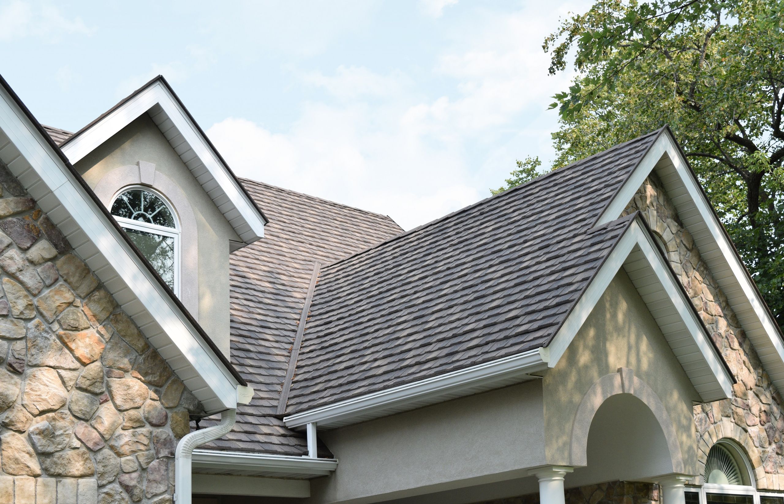 Close-up of a house roof with stone-coated metal shingles in a gray tone, featuring gable ends, arched window, and stone exterior walls, surrounded by trees under a partly cloudy sky