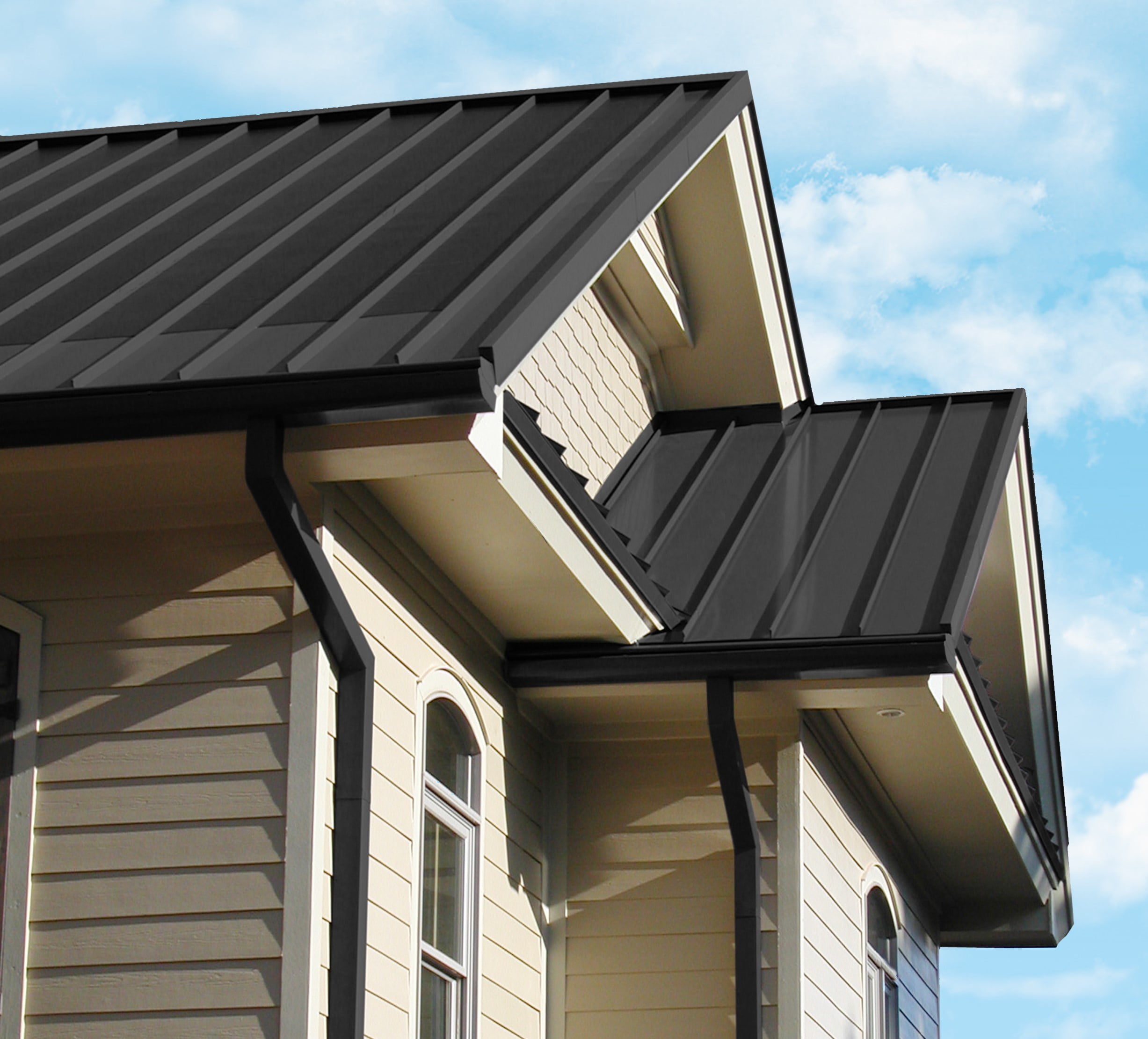 Seamless Metal Roof Close-up view of a house with a dark gray seamless metal roof, light beige siding, and dark gray gutters against a blue sky with scattered clouds.