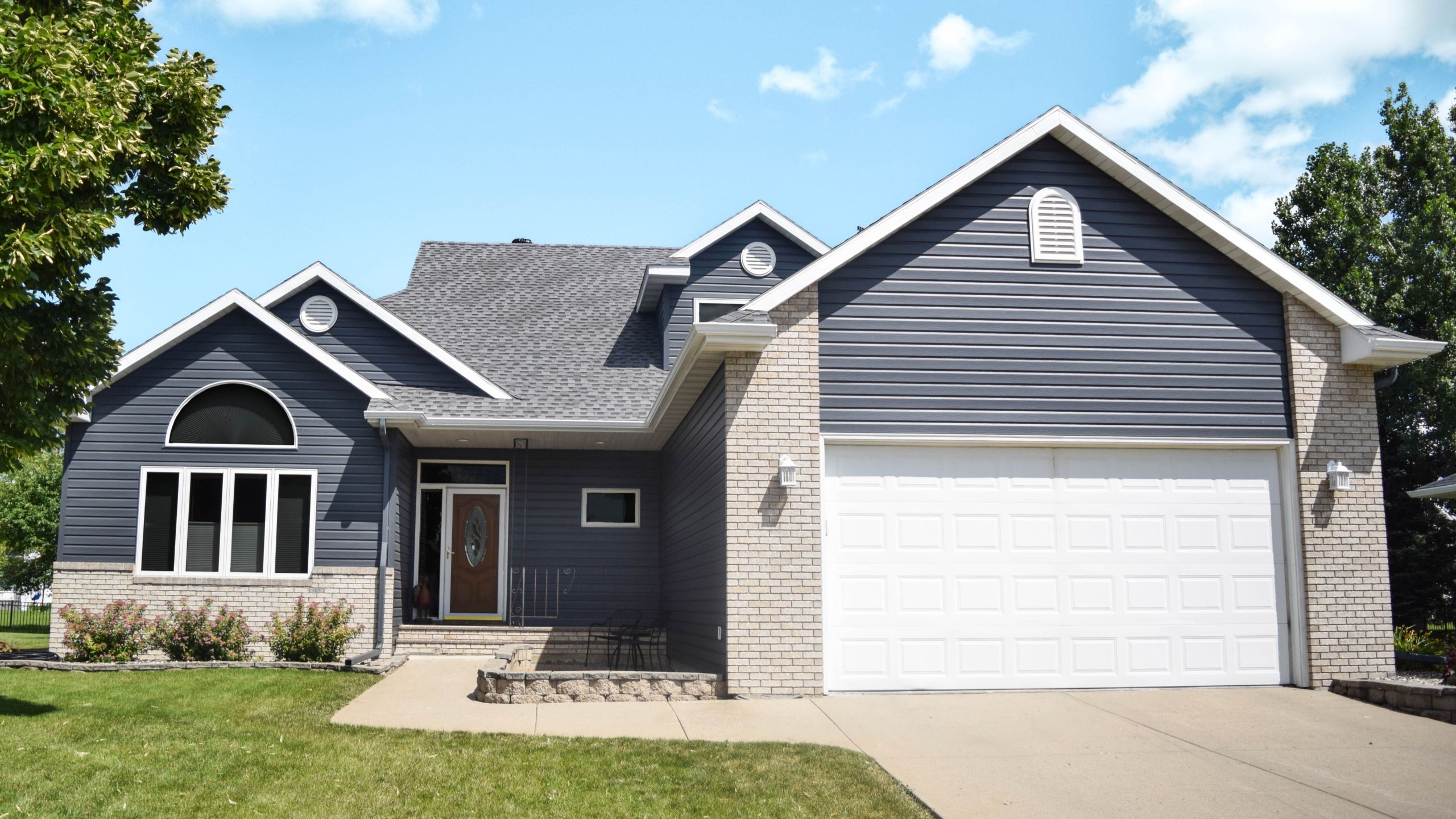 Home exterior with dark gray-blue seamless steel siding, white trim and garage door, and brick accents under a blue sky.