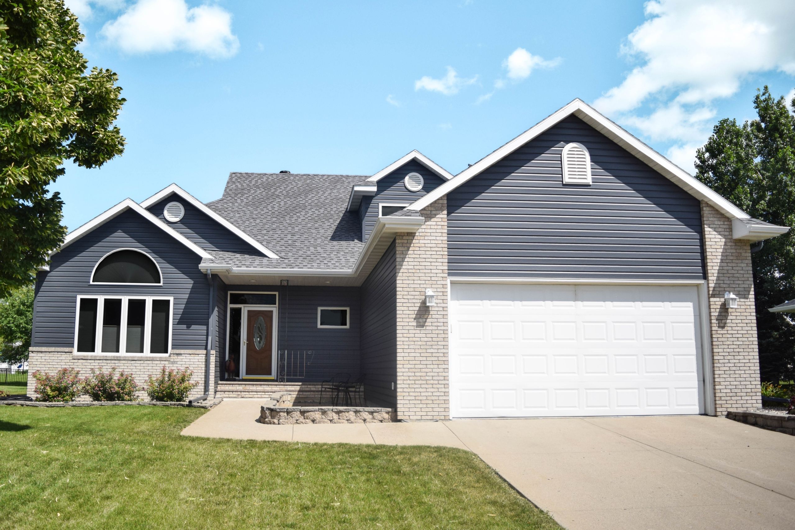 Home exterior with dark gray-blue seamless steel siding, white trim and garage door, and brick accents under a blue sky.