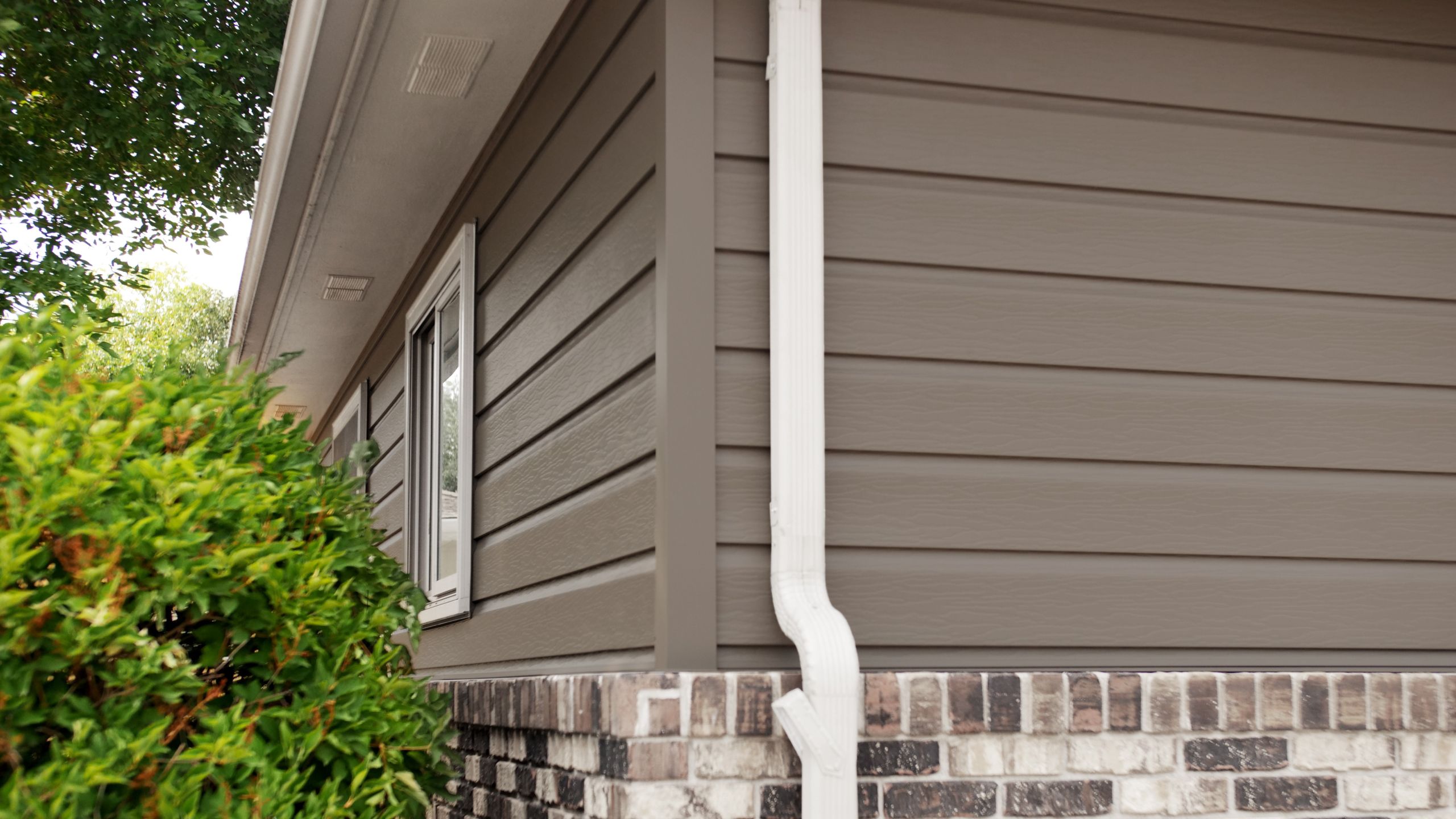 Close-up of a home exterior with brown seamless steel siding, white trim and gutter, and brick accents next to green shrubs.